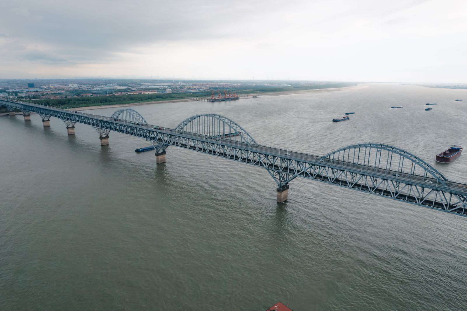 Aerial view of a long steel arch bridge crossing a wide stretch of river with cargo boats below