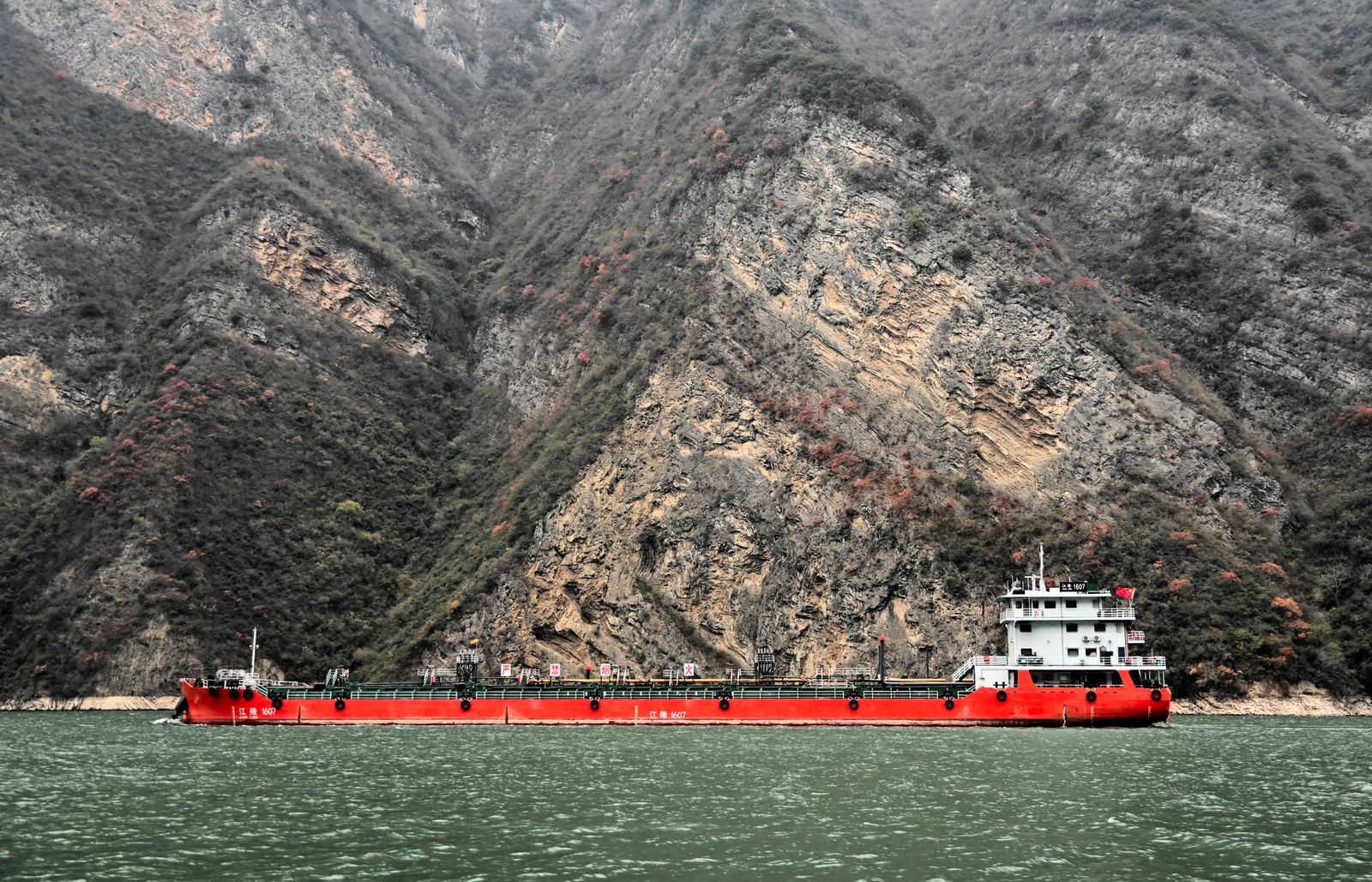 A red Chinese-flagged river tanker passing below steep, folded rock walls in a narrow gorge, a reminder that cruise ships share the Yangtze with working traffic