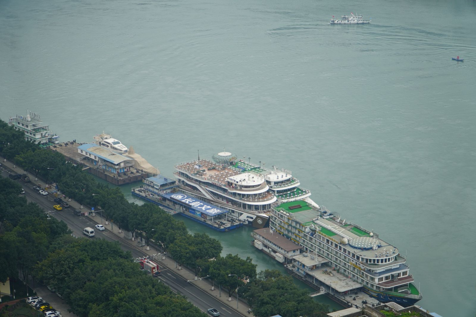 Aerial view of large multi-deck river cruise ships moored at floating piers along a broad, tree-lined city riverfront, the kind of ships compared on this page