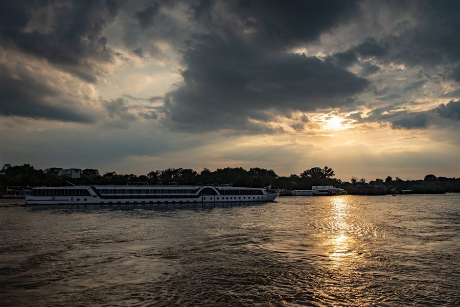 A long white passenger riverboat sailing past a wooded bank under dramatic sunset clouds
